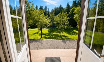 An open terrace door overlooking a sunny garden. Green trees and a clear blue sky create an inviting atmosphere.