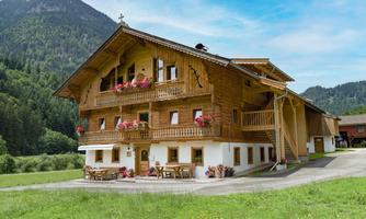 A traditional wooden chalet with balconies and flower boxes. The surroundings are green and surrounded by mountains.