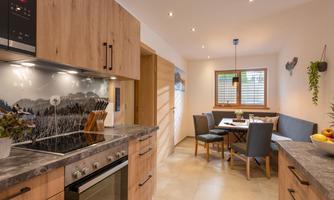 A modern kitchen with wooden cabinets and a cooktop. In the background, there is a dining area with a table and chairs.