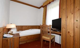 A simple room with wooden walls, a single bed, and a desk. A television is on the desk, and the window lets in a lot of light.