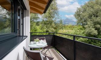 A beautiful balcony with a table and two chairs, surrounded by trees and green countryside. In the background, mountains and a clear sky are visible.