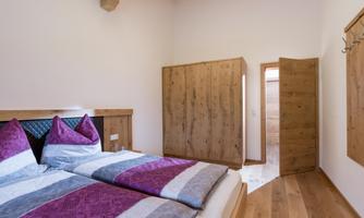 A cozy bedroom with a double bed and wooden furniture. The walls are simple, and there is an entrance to a bathroom.