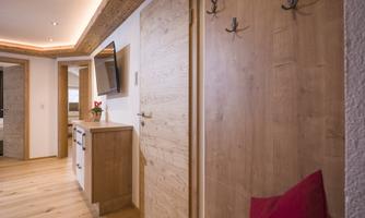 A modern hallway with wooden paneling and a red cushion. The wall displays coat hooks, and in the background, there is a door as well as a television.