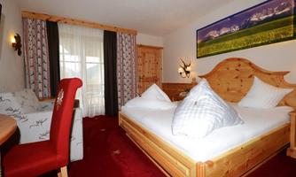 A cozy room with a wooden double bed and two large pillows. There is also a small table, an armchair, and large windows that let in plenty of light.