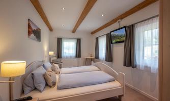 A bright bedroom with two beds, cozy lighting, and modern furniture. The room has large windows with curtains and a television on the wall.