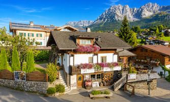 A picturesque alpine house with colorful flower balconies, surrounded by green meadows and striking mountains in the background. The sun is shining and the sky is clear.