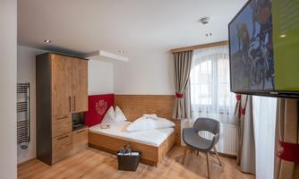 A cozy room with a single bed, a modern television, and a chair. The decor is bright and friendly, with wooden furniture and a large window.