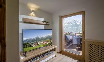 A cozy living space with a television and a view of the mountains through a large window. The room is bright and modernly furnished.