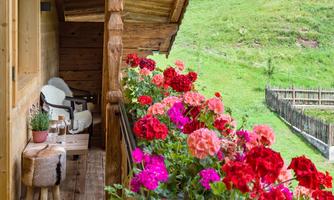 A balcony with colorful flowers and two cozy chairs. In the background, a green meadow can be seen.