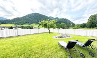 A green meadow with two loungers and a view of gentle hills. In the background, mountains and a blue sky can be seen.
