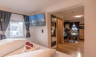 A modern room with a double bed and a large window. In the corner, there is a flat-screen TV and an adjacent dining area.