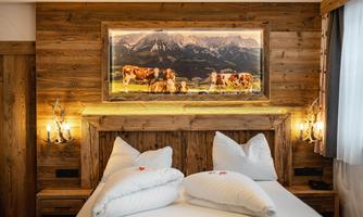 A cozy bedroom with a wooden bed and white pillows. A picture of cows in a mountainous landscape hangs on the wall.