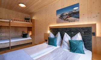 A cozy bedroom with a modern wood design. There are two beds, a bunk bed, and decorative cushions in a relaxing color scheme.