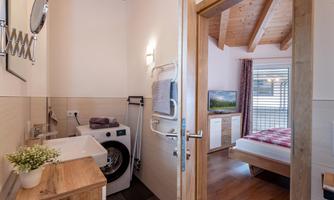 A modern bathroom with a washing machine and wood details. In the background, there is a bedroom with a colorful bed and a television.