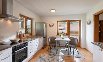 A modern kitchen with a large window and a bright wooden ceiling. The room is furnished with a dining table and elegant chairs.