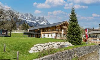 A cozy mountain cabin surrounded by green meadows and trees. In the background, impressive mountains and a clear sky can be seen.