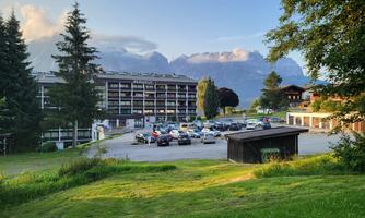 A large building in the Alps with parking spaces in the foreground. The surroundings are green and surrounded by mountains.