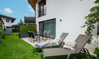 A cozy outdoor area with loungers and a table in the garden. The house has large windows and a modern facade.