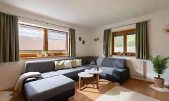 A cozy living room with a gray sofa and a small table. Large windows let in plenty of daylight, and plants add a fresh touch to the environment.