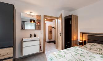 A modern bedroom with a double bed and a minimalist wardrobe. In the background, a bathroom with a sauna is visible.