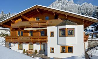 A chalet-shaped wooden house with several balconies stands in the snow. Snow-covered mountains can be seen in the background.