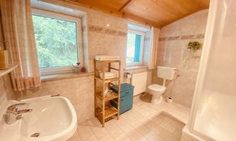 A bright bathroom with a window, a sink, and a shower cabin. The room also has a shelf with towels and personal care products.