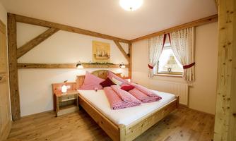 A cozy bedroom with a large wooden bed and colorful pillows. The walls are decorated with beautiful wooden beams and there is a window with curtains.