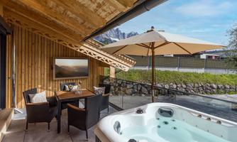 A cozy outdoor area with a hot tub, a table, and chairs. In the background, the mountains and a blue sky are visible.