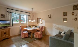 A cozy dining area with a table and two chairs, surrounded by warm colors. A sofa is next to the table and a television is visible in the background.