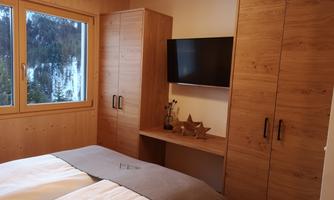A cozy bedroom with two beds and wooden wardrobes. A television hangs on the wall and there is a window with a view of nature.