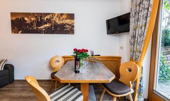 A cozy dining area with a wooden table and wooden chairs. On the table is a vase with red roses, and in the background, there is a television and a landscape painting.
