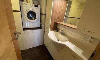 A small bathroom with a washing machine and a modern sink. The walls are brightly tiled and there is a wooden shelf.