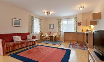 A bright, modern furnished apartment with a red sofa and a dining area. The windows let in plenty of light and the floor is covered with a carpet.
