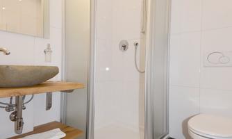 A modern bathroom with a glass shower and a simple sink. The floor is tiled with dark tiles.