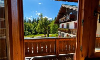 A view from the balcony of a picturesque house surrounded by trees and meadows. The sky is blue with some clouds, and it seems to be sunny.