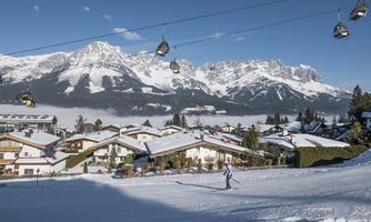 A snow-covered landscape with a skier and picturesque mountain peaks in the background. The houses are visible in the foreground, while the cable cars glide over the scene.