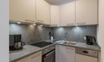 A modern kitchen with light cabinets and a gray countertop. It is equipped with a cooktop, an oven, and a coffee machine.