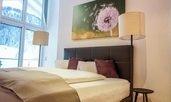 A modern bedroom with a large bed and soft pillows. A picture of a dandelion hangs on the wall, and there are two floor lamps.