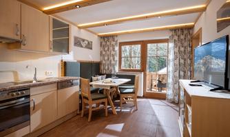 A modern kitchen with wood and glass design. The room has a dining table and large windows that offer a view outside.
