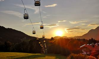 A cable car travels over a green meadow towards the sunset. In the background, there are mountains and a beautiful, slightly cloudy sky.