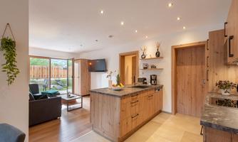 A modern kitchen with wooden cabinets and a stone countertop. The open space leads to a cozy living area with access to the garden.