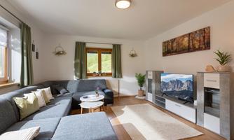 A bright living room with a gray sofa, a table, and a television.  
Large windows let in plenty of daylight and the decoration is minimalist and modern.