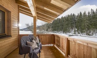 A wooden balcony with a cozy armchair and a view of snow-covered trees. The sun is shining and the sky is clear.