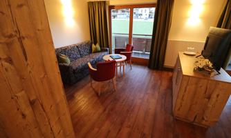 A cozy room with wooden floors, a sofa, and a table. Through the window, you can see a balcony that lets in plenty of daylight.