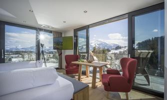 A modern room with large windows and a stunning view of the mountains. Red armchairs and a table invite you to relax.