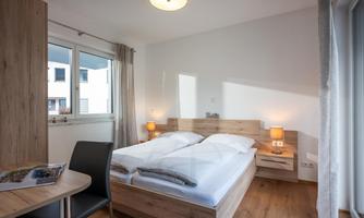 A modern bedroom with a double bed and wooden furniture. Large windows provide plenty of daylight.