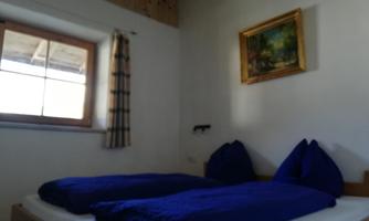 A cozy bedroom with a window and a colorful painting on the wall. Two beds with blue bedspreads are ready.
