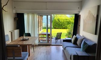 A cozy living space with a dining area and a sofa. Large windows provide a view of the outdoor garden.