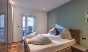 A modern bedroom with a double bed and large pillows. The walls are painted in soft colors, and there are large windows that let in plenty of light.