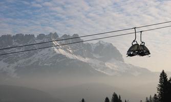A cable car glides over a mountainous landscape. In the background, snow-covered peaks and a cloudy sky are visible.